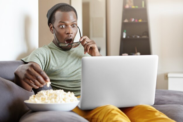 Sorprendido hombre africano sentado en el sofá de su casa, comiendo palomitas de maíz y viendo un emocionante programa de televisión en línea en la computadora portátil o sorprendido con el final de la serie de detectives, con la boca abierta