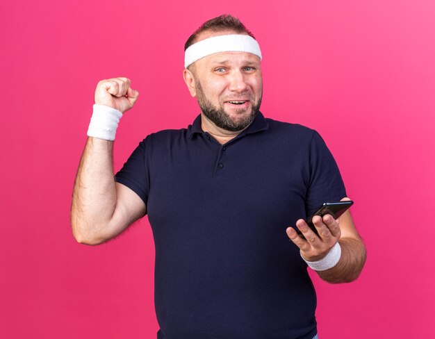 Sorprendido hombre deportivo eslavo adulto con diadema y muñequeras sosteniendo el teléfono y manteniendo el puño hacia arriba aislado en la pared rosa con espacio de copia