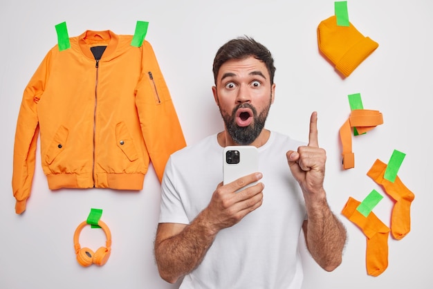 Sorprendido hombre guapo con barba gruesa mantiene el dedo índice levantado tiene una idea brillante sostiene el teléfono móvil para poses de comunicación en línea contra fondo blanco ropa y accesorios enyesados