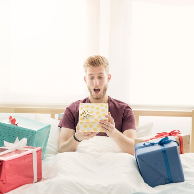 Sorprendido joven apertura caja de regalo sentado en la cama