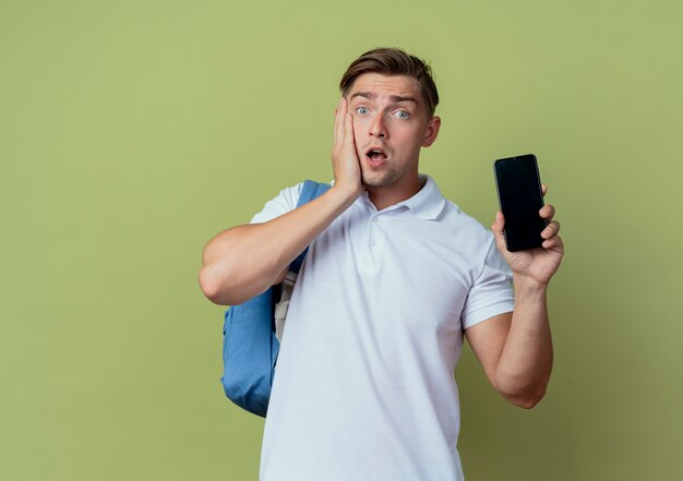 Sorprendido joven apuesto estudiante vistiendo bolso trasero sosteniendo el teléfono y poniendo la mano en la mejilla aislada en verde oliva