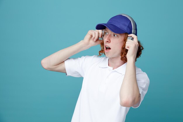 Sorprendido joven apuesto con gorra usando auriculares aislados de fondo azul
