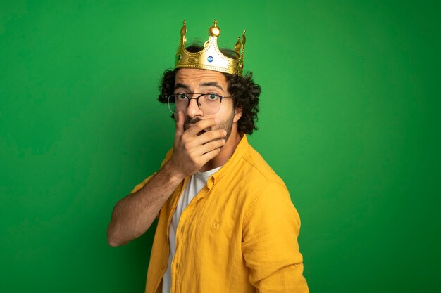 Sorprendido joven apuesto hombre caucásico con gafas y corona de pie en la vista de perfil mirando a la cámara manteniendo la mano en la boca aislada sobre fondo verde con espacio de copia