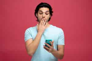 Foto gratuita sorprendido joven apuesto manteniendo la mano en la boca sosteniendo y mirando el teléfono móvil aislado en el fondo rojo