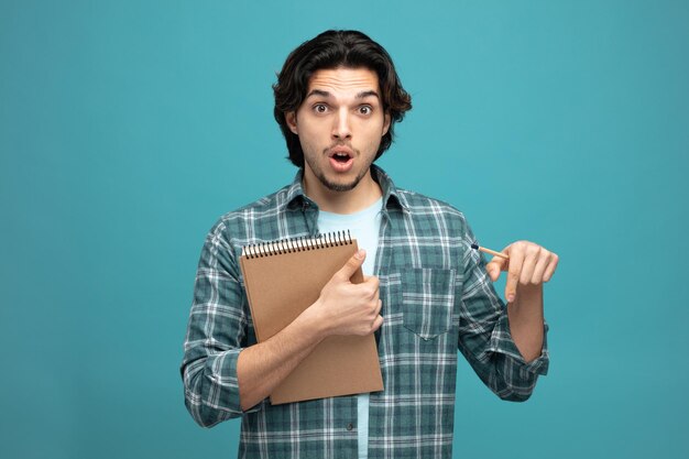 Sorprendido joven apuesto mirando a la cámara apuntando hacia abajo con lápiz y bloc de notas en las manos aisladas en el fondo azul