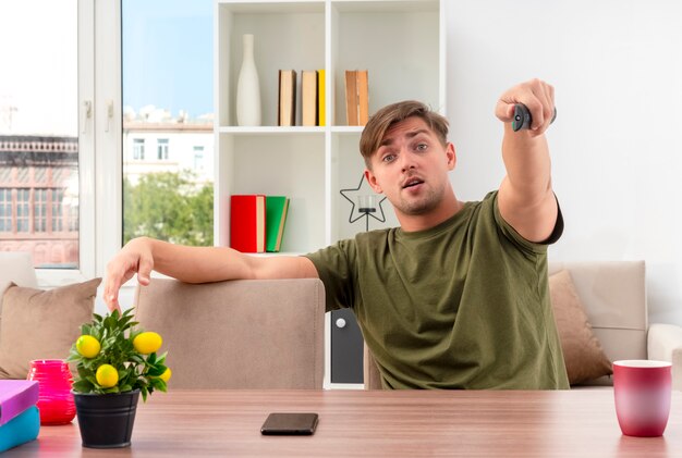 Sorprendido joven apuesto rubio se sienta a la mesa sosteniendo el control remoto de la televisión mirando a la cámara dentro de la sala de estar