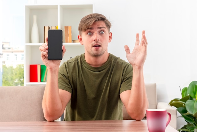 Sorprendido joven apuesto rubio se sienta a la mesa con una taza sosteniendo el teléfono y levantando la mano dentro de la sala de estar