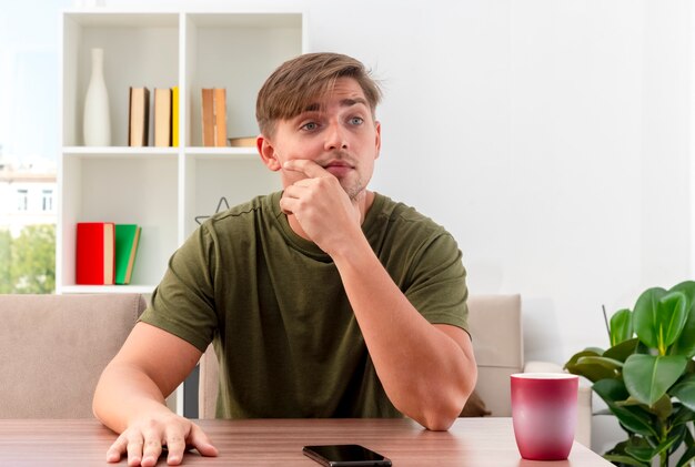 Sorprendido joven apuesto rubio se sienta a la mesa con el teléfono y la taza sosteniendo la barbilla y mirando de lado dentro de la sala de estar