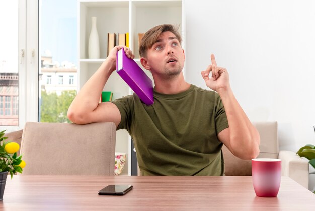 Sorprendido joven apuesto rubio se sienta a la mesa con el teléfono y la taza sosteniendo el libro en el hombro mirando y apuntando hacia arriba dentro de la sala de estar