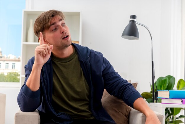 Sorprendido joven apuesto rubio se sienta en un sillón apunta hacia arriba mirando al lado dentro de la sala de estar