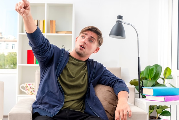 Sorprendido joven apuesto rubio se sienta en un sillón mirando y apuntando a un lado dentro de la sala de estar