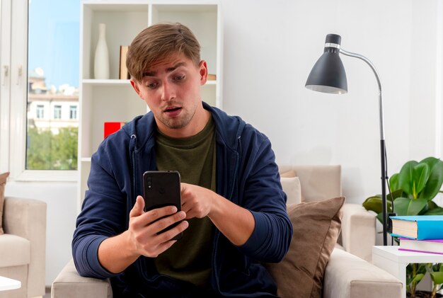 Sorprendido joven apuesto rubio se sienta en un sillón sosteniendo y mirando el teléfono dentro de la sala de estar