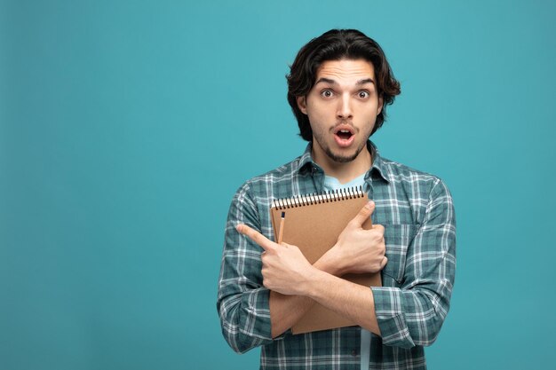 Sorprendido joven apuesto sosteniendo lápiz y bloc de notas mirando a la cámara apuntando al lado aislado en fondo azul con espacio para copiar