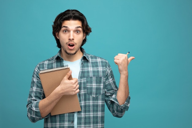 Sorprendido joven apuesto sosteniendo lápiz y bloc de notas mirando a la cámara apuntando al lado aislado en fondo azul con espacio para copiar