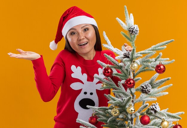 Sorprendido joven asiática vistiendo gorro de navidad con suéter de pie cerca del árbol de navidad extendiendo la mano aislada sobre fondo naranja