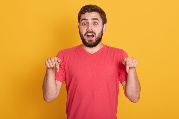 sorprendido joven asustado que tiene miedo de algo, apuntando hacia abajo con los dedos delanteros, después de haber sorprendido y asombrado la expresión facial, posando aislado en amarillo