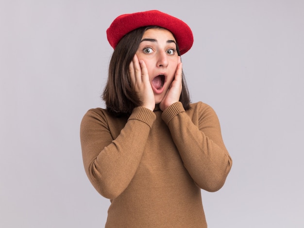 Sorprendido joven bastante caucásica con sombrero de boina pone las manos en la cara