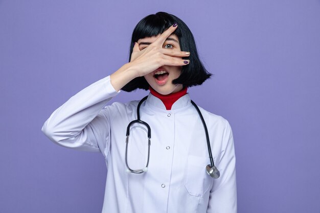 Sorprendido joven bastante caucásica en uniforme médico con estetoscopio cubriendo su rostro con la mano y mirando al frente a través de los dedos