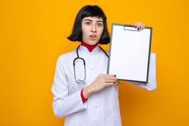 Sorprendido joven bastante caucásica en uniforme médico con estetoscopio sosteniendo portapapeles