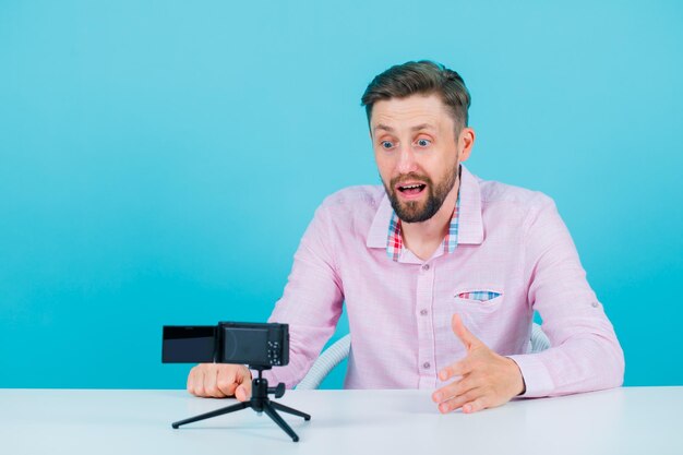 Sorprendido joven bloguero está posando para su mini cámara con fondo azul