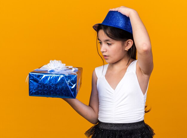 Sorprendido joven caucásica con gorro de fiesta azul poniendo la mano en el sombrero y mirando la caja de regalo aislada en la pared naranja con espacio de copia