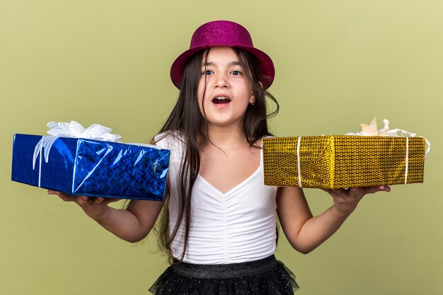 Sorprendido joven caucásica con gorro de fiesta púrpura sosteniendo una caja de regalo en cada mano aislado en la pared verde oliva con espacio de copia