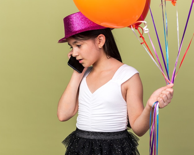 Foto gratuita sorprendido joven caucásica con gorro de fiesta púrpura sosteniendo globos de helio hablando por teléfono aislado en la pared verde oliva con espacio de copia