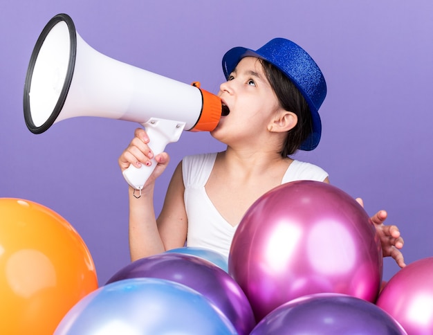 Sorprendido joven caucásica con sombrero de fiesta azul hablando por el altavoz mirando hacia arriba de pie con globos de helio aislado en la pared púrpura con espacio de copia