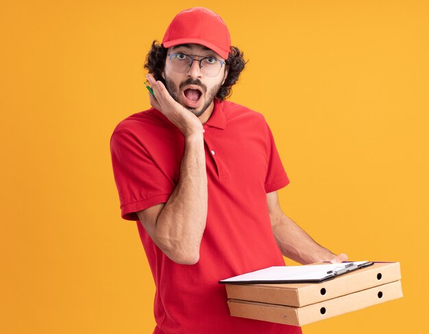 Sorprendido joven caucásico repartidor en uniforme rojo y gorra con gafas sosteniendo paquetes de pizza lápiz portapapeles mirando al frente manteniendo la mano en la cara