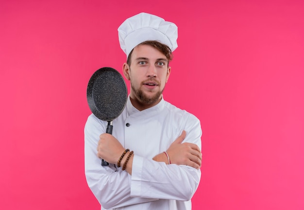 Foto gratuita un sorprendido joven chef barbudo en uniforme blanco con gorro de cocinero sosteniendo una sartén mientras mira en una pared rosa