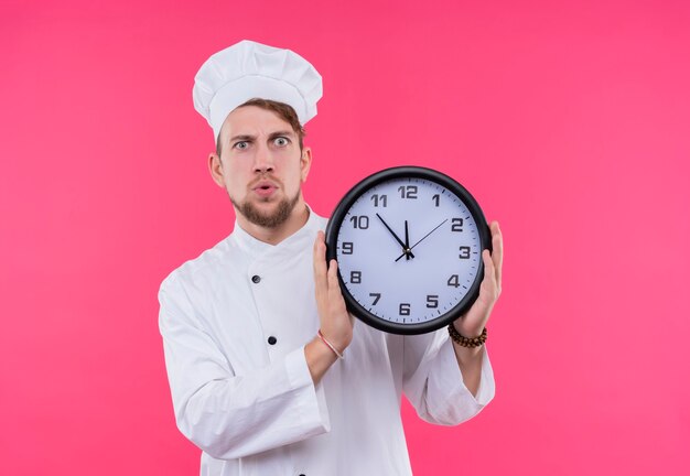 Un sorprendido joven chef barbudo con uniforme blanco que muestra el tiempo mientras sostiene el reloj de pared en una pared rosa