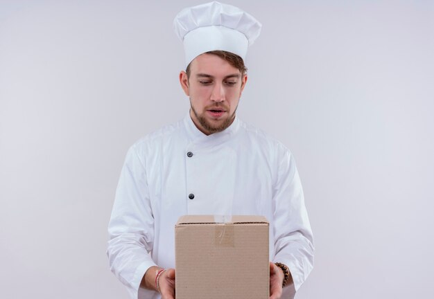 Un sorprendido joven chef barbudo en uniforme blanco sosteniendo la caja de entrega y mirándola en una pared blanca