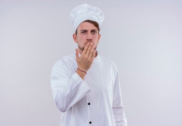 Un sorprendido joven chef barbudo vestido con uniforme de cocina blanco y sombrero manteniendo la mano en la boca mientras mira en una pared blanca