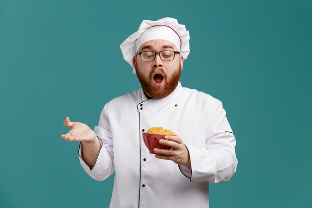 Sorprendido joven chef masculino con uniforme de anteojos y gorra sosteniendo y mirando el tazón de macarrones mostrando la mano vacía aislada en el fondo azul
