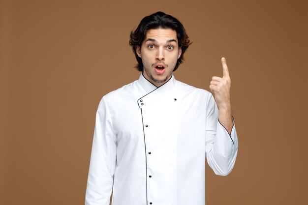 sorprendido joven chef masculino vistiendo uniforme mirando a la cámara apuntando hacia arriba aislado sobre fondo marrón