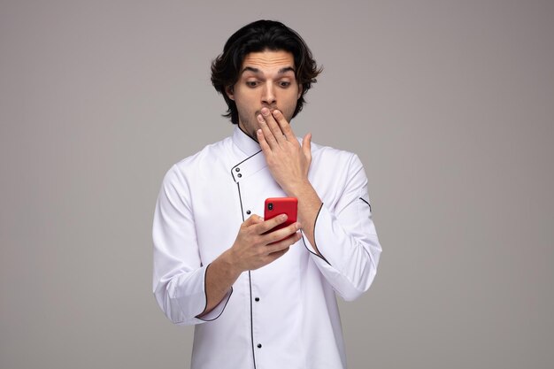 Sorprendido joven chef uniformado sosteniendo un teléfono móvil mirándolo manteniendo la mano en la boca aislada de fondo blanco