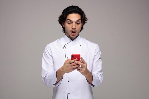 Sorprendido joven chef uniformado usando su teléfono móvil aislado de fondo blanco