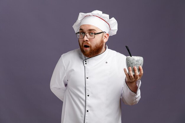 Sorprendido joven chef con uniforme de anteojos y gorra que muestra un tazón con una cuchara mirando a la cámara mientras mantiene la mano detrás de la espalda aislada en un fondo morado