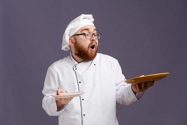 Sorprendido joven chef con uniforme de anteojos y gorra sosteniendo platos vacíos mirando uno aislado de fondo morado