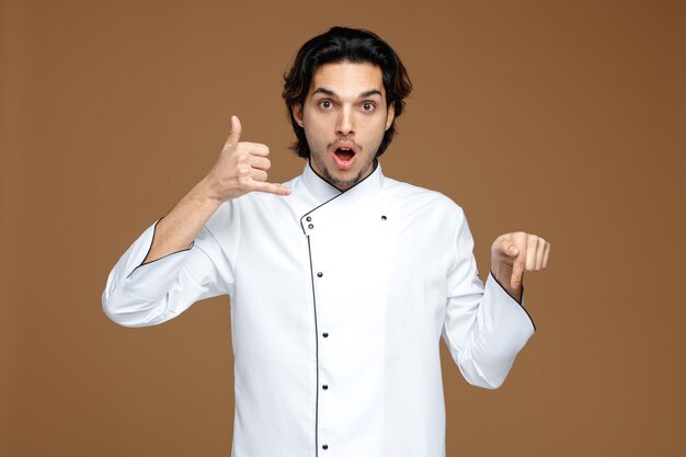 Sorprendido joven chef con uniforme que muestra un gesto de llamada apuntando hacia abajo aislado en un fondo marrón