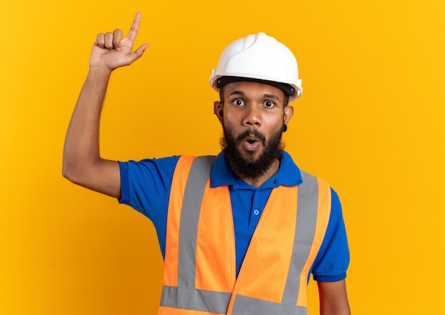 Sorprendido joven constructor afroamericano en uniforme con casco de seguridad apuntando hacia arriba aislado sobre fondo naranja con espacio de copia