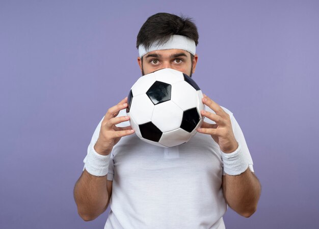 Sorprendido joven deportivo vistiendo diadema y muñequera cubrió la cara con bola