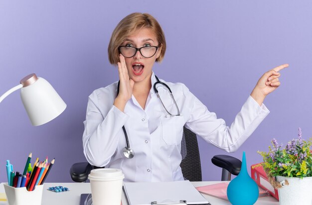 Sorprendido joven doctora vistiendo bata médica con estetoscopio y gafas se sienta a la mesa con herramientas médicas llamando a alguien apunta al lado aislado sobre fondo azul.