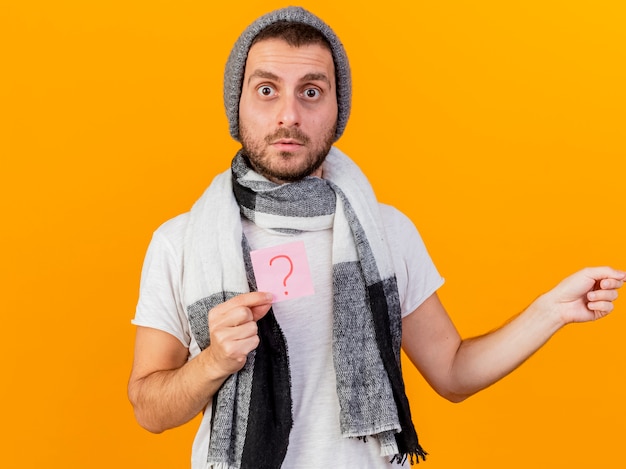 Sorprendido joven enfermo vestido con gorro de invierno y bufanda con nota de signo de interrogación y puntos en el lado aislado sobre fondo amarillo