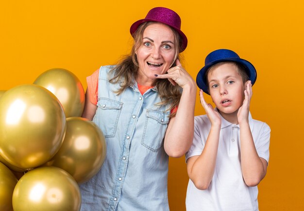 Sorprendido joven eslavo con sombrero de fiesta azul de pie levantando las manos con su madre con sombrero de fiesta púrpura sosteniendo globos de helio aislados en la pared naranja con espacio de copia