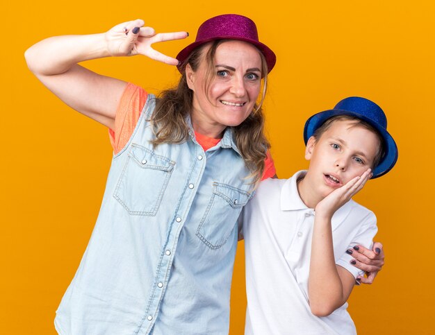 Sorprendido joven eslavo con sombrero de fiesta azul poniendo la mano en la cara y de pie con su madre con sombrero de fiesta púrpura gesticulando signo de victoria aislado en la pared naranja con espacio de copia