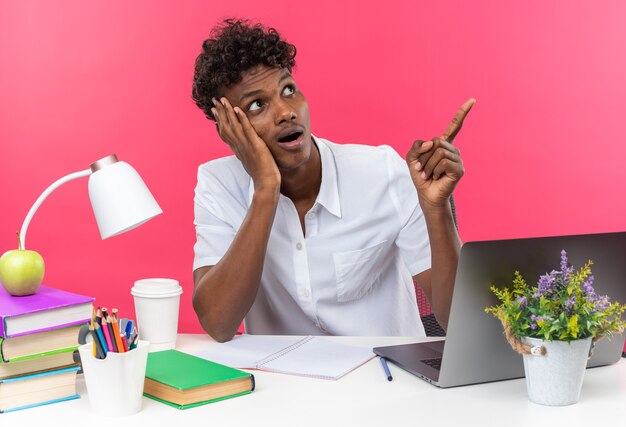 Sorprendido joven estudiante afroamericano sentado en el escritorio con herramientas escolares poniendo la mano en la cara mirando y apuntando al lado aislado en la pared rosa