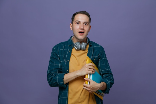 Sorprendido joven estudiante masculino usando audífonos alrededor del cuello sosteniendo herramientas de estudio bajo el brazo mirando al lado aislado en un fondo morado