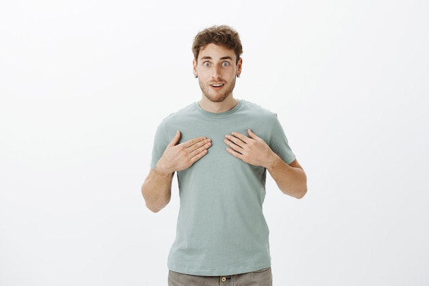 Sorprendido joven feliz sintiéndose emocionado mientras está de pie, apuntando a sí mismo, sonriendo y sin palabras