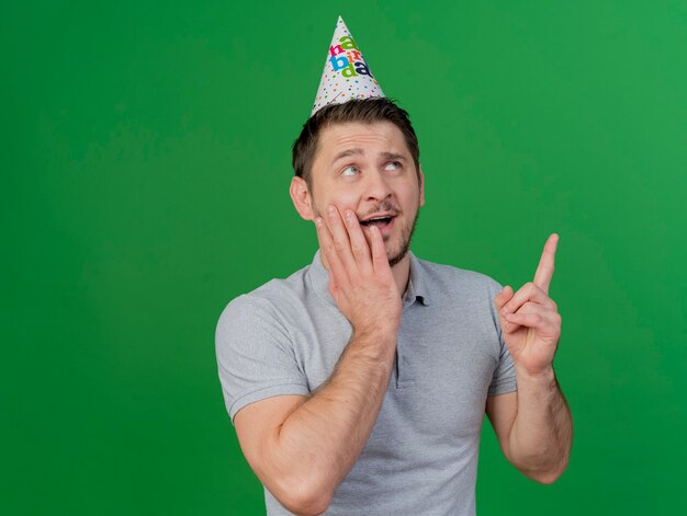 Foto gratuita sorprendido joven fiestero vistiendo gorro de cumpleaños poniendo la mano en los puntos de la mejilla en aislado en verde